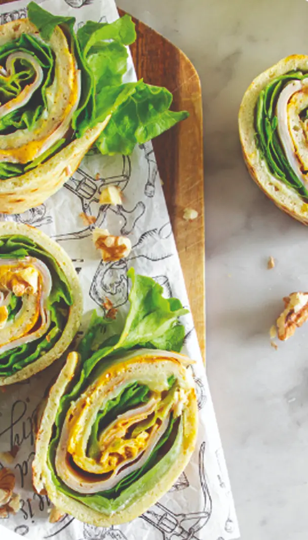 Rotoli di piadina con prosciutto, lattuga e formaggio, appoggiati su carta da forno con alcune noci sparse sul tavolo.