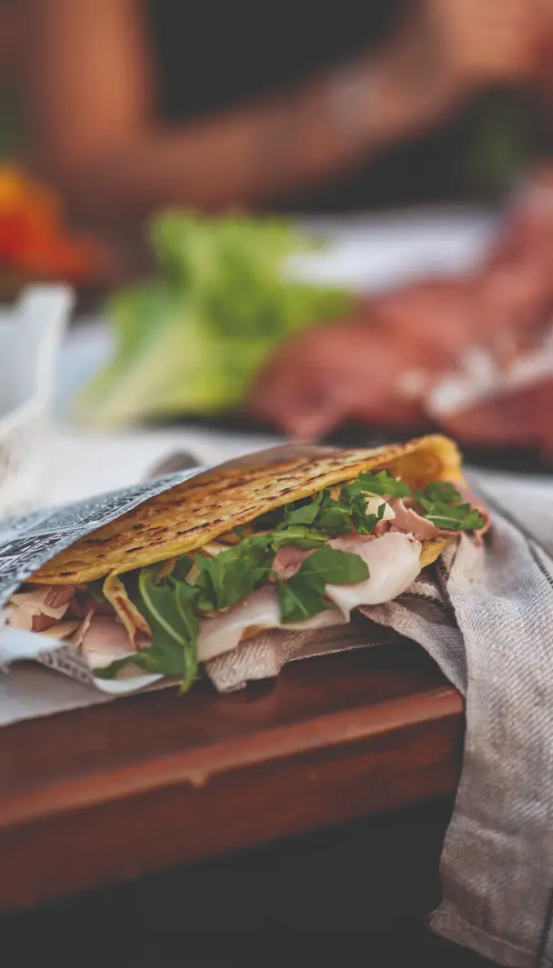 Crostolo ripieno di prosciutto e rucola, appoggiato su un tovagliolo di stoffa con verdure sullo sfondo.