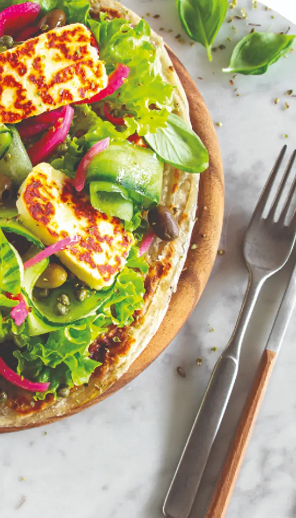Piadina sfogliata con formaggio halloumi grigliato, lattuga, cetrioli, cipolla rossa e olive, accompagnata da forchetta e coltello su un piatto di legno.