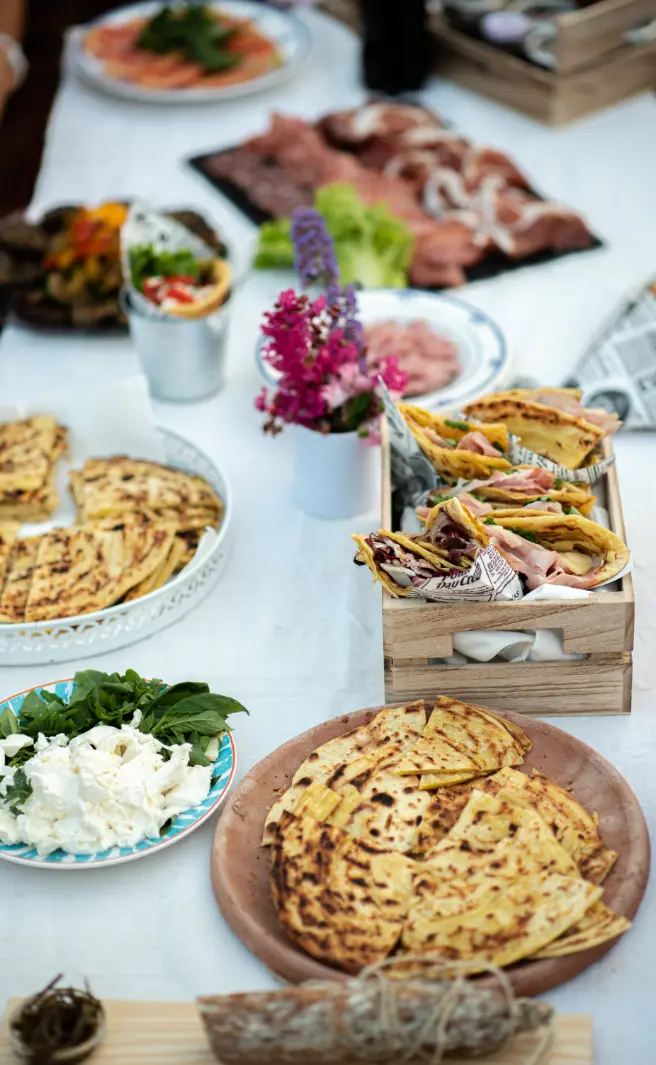 Un tavolo apparecchiato con piadine sfogliate, affettati, insalata, fiori freschi e formaggio, in un ambiente rustico e accogliente.