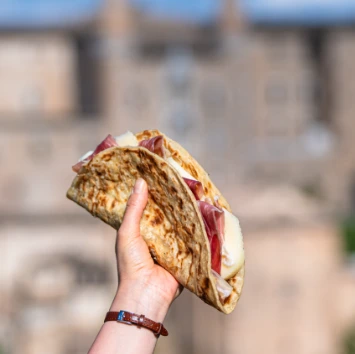 Una mano che tiene una crescia sfogliata farcita con prosciutto e formaggio, con il paesaggio di Urbino sullo sfondo.