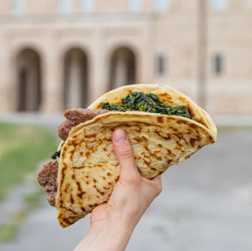 Una mano che tiene un crostolo farcito con salsiccia e verdure, con uno sfondo che mostra un edificio storico.