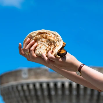 Una mano che tiene una piadina sfogliata farcita con ingredienti, con il cielo azzurro e la rocca di Senigallia sullo sfondo.