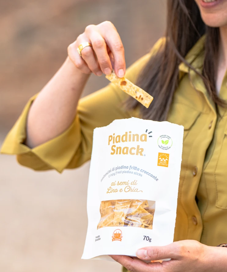 Una donna che estrae un bastoncino di Piadina Snack ai semi di Lino e Chia dal pacchetto, mentre lo tiene in mano. Il pacchetto è visibile, con la scritta 'Piadina Snack' e il logo.